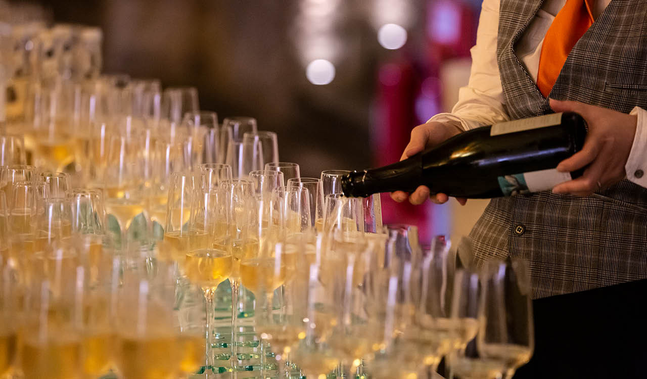 Sparkling wine is poured into glasses by someone wearing an orange tie and tweed waistcoat.