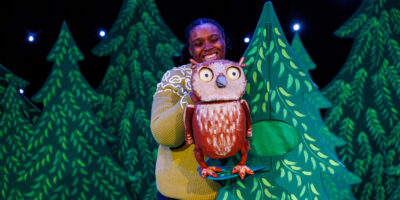 An actor operates a puppet of an owl, as if the owl is sitting in a tree.