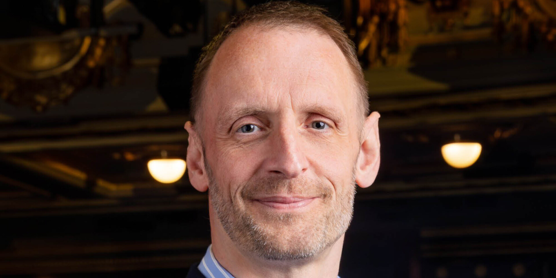 A close up headshot of Darren Atkins, looking directly at the camera with a soft smile. His shirt has a blue collar with white stripes, which is just visible in frame.