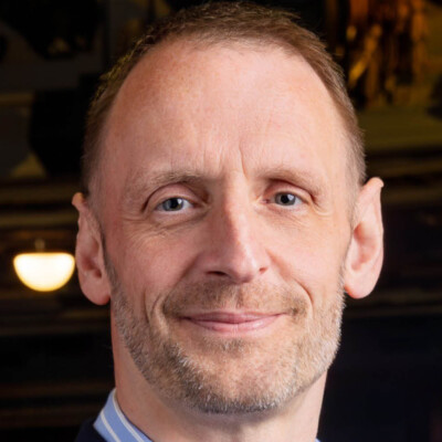A close up headshot of Darren Atkins, looking directly at the camera with a soft smile. His shirt has a blue collar with white stripes, which is just visible in frame.