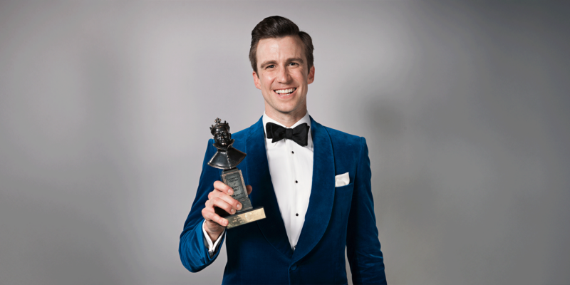 Gavin Creel, dressed in a blue velvet suit and bow tie, holds his Olivier Award while smiling at the camera.