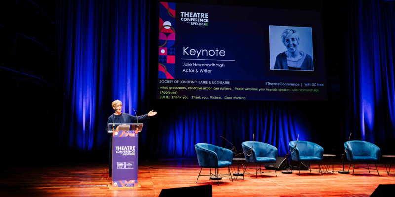 Julie Hesmondhalgh stood behind a podium delivering a speech next to a big screen with her photo and 'Keynote' written.