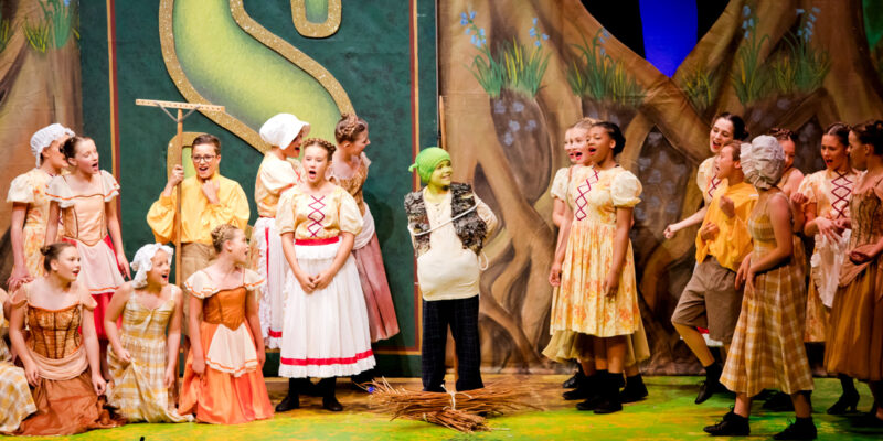Children perform in Shrek The Musical. One child in the centre is dressed in a green hat with green facepaint, and two groups of children either side wear medieval-style costumes and are singing.