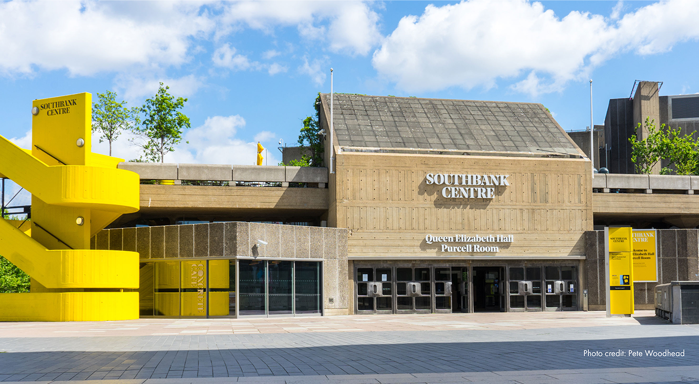 Photo of the Queen Elizabeth Hall at the Southbank Centre. Brutalist architecture with a bright yellow curved staircase on the left.
