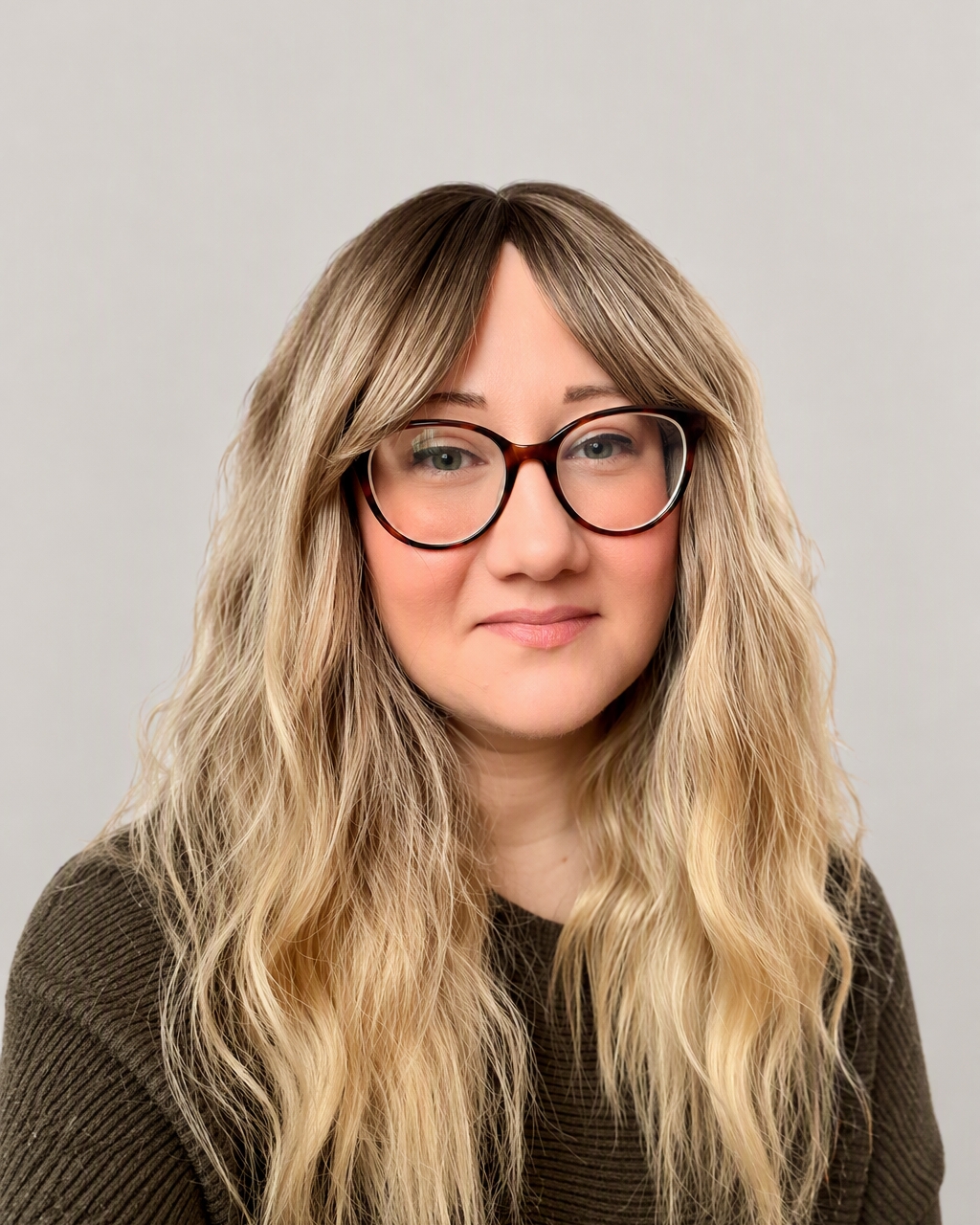 A headshot photo of Becci. She is a caucasion woman with blonde, wavy hair, black round glasses and is wearing a dark green jumper.